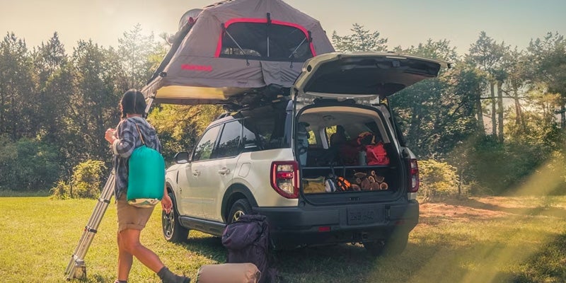 Man walking away from a white 2024 Ford Bronco Sport that is equipped with a tent on the roof rack and other camping gear in the trunk with a wooded landscape in the background